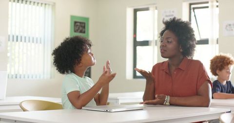 African american teacher guiding young boy with gestures at sunlit classroom table