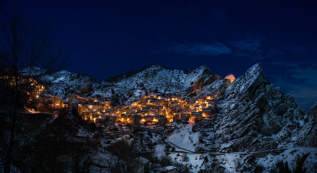 Snow-covered winter village nestled in mountains, beautifully lit at night. Great for travel promotions, winter tourism, holiday postcards, nature photography, serene landscape visuals.