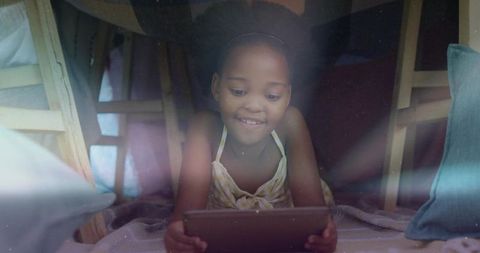 Child watching tablet inside cozy blanket fort with pillows, soft glow, warm indoor play