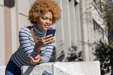 African american woman leaning on concrete ledge checking smartphone in sunlit cityscape