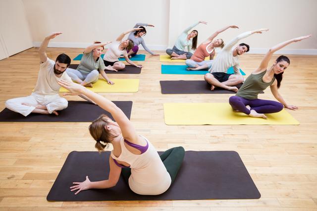 Group of people practicing yoga stretching exercises on mats in a fitness studio. Ideal for use in articles or advertisements related to fitness, wellness, yoga classes, group workouts, and healthy lifestyles.