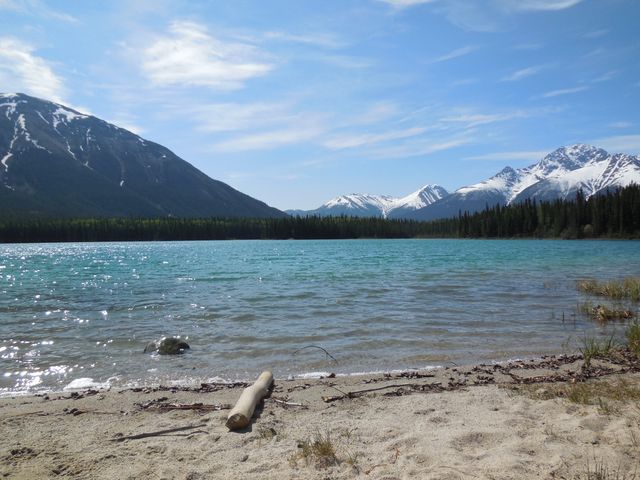 Mountain lake with clear waters and snowy peaks in the background. Sandy shoreline in foreground, with blue sky and a few clouds. Perfect for travel brochures, nature calendars, outdoor adventure blogs, peaceful retreat advertisements.