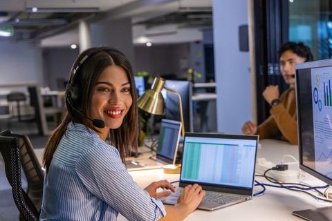 Smiling support agent typing on laptop with headset in modern open office