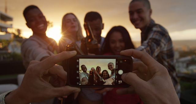 Friends are celebrating by taking group selfie on an urban rooftop during sunset. This scene captures the essence of togetherness, joy, and friendship among diverse young adults. Ideal for promotions about social events, friendship, smartphone usage, or urban lifestyle. Great for blogs, social media campaigns, or advertisements targeting youthful and energetic audiences.