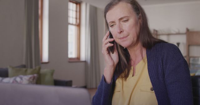 Mature woman sitting at home, talking on a mobile phone while using a laptop. Ideal for illustrating concepts of remote work, telecommuting, work-life balance, effective communication, and professional women. Can be used in articles, blogs, and advertisements related to remote working trends, technology use, and productivity at home.