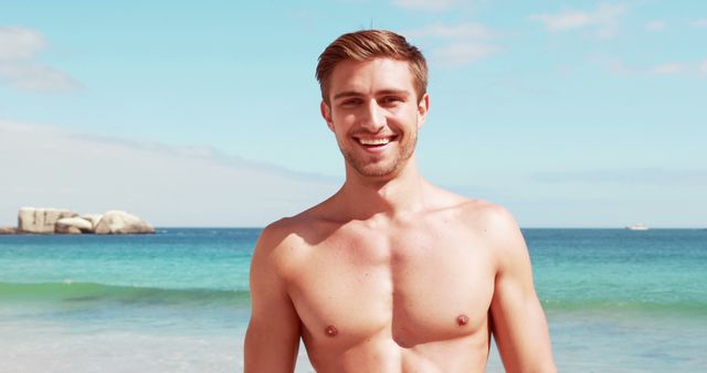 A young Caucasian man smiles while standing shirtless on a sunny beach, with copy space - Download Free Stock Photos Pikwizard.com