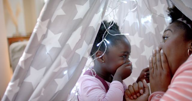 Mother and daughter are sharing a fun and playful moment inside a star-patterned play tent adorned with fairy lights. The scene suggests a warm, intimate family bonding time filled with laughter and joy. This image can be used for articles and advertisements celebrating parenthood, childhood memories, family activities, or indoor playtime.