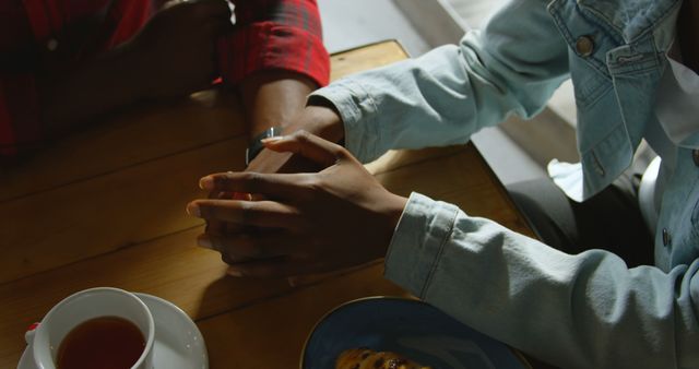 Two individuals are holding hands across a table in a gesture of comfort or intimacy, with copy space. A warm beverage and a pastry on the table suggest a cozy, supportive atmosphere.