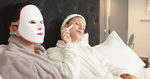 Couple relaxing in bed wearing led facial mask and using skincare device during home evening spa