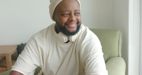 African man smiling and relaxing in green armchair wearing cream beanie and headphones