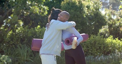 Sunlit park friends hugging after outdoor yoga session, african american duo holding mats