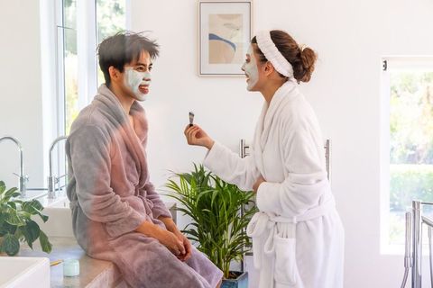 Couple enjoying at-home spa night applying facial masks in sunlit bathroom with plants and robes