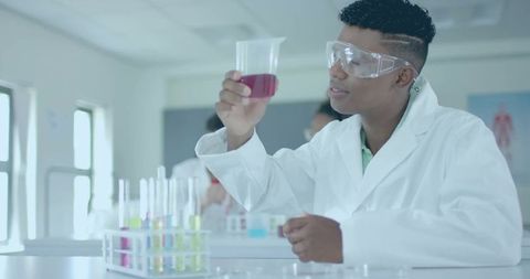 Teen examining red solution in high school chemistry lab wearing goggles and lab coat