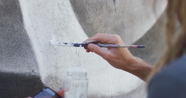 This close-up captures an artist working on a mural, highlighting the use of a paintbrush and white paint on a textured wall. Ideal for websites and blogs focused on street art, creative processes, urban culture, and artistic expression. Can also be used for articles or features on muralists and their techniques.