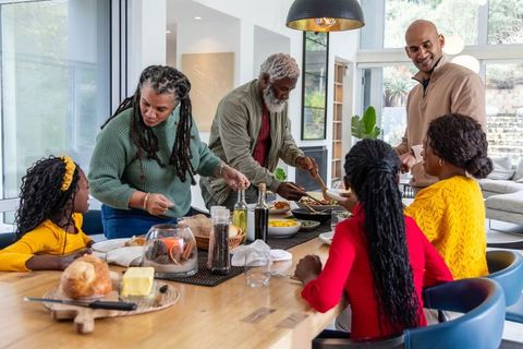 Multigenerational black family serving homemade meal around sunlit open-plan dining table