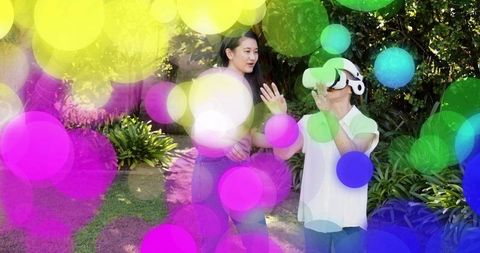 Younger woman guiding senior wearing vr headset in sunny backyard with colorful bokeh