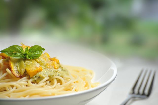 Freshly cooked spaghetti topped with creamy pesto sauce and roasted vegetables served in a white bowl with a basil garnish. Ideal for use in food blogs, Italian cuisine websites, recipe books, cooking magazines, and restaurant menus showcasing gourmet vegetarian or vegan dishes.