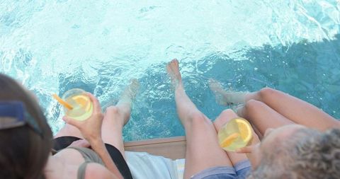 Three women dipping feet in pool while holding citrus drinks poolside summer relaxation