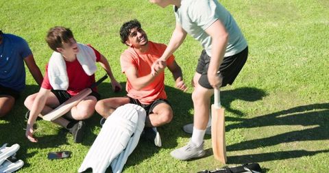 Cricket teammates exchanging handshake after match on sunny grassy outfield with bats and pads