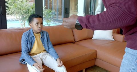 Concerned african american father gesturing while son sits on leather sectional near pool