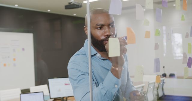 Businessman concentrating while adding ideas, plans, and strategies on sticky notes on glass wall in modern office. Ideal for depicting brainstorming sessions, workplace creativity, project planning, entrepreneurial strategies, and team collaboration.