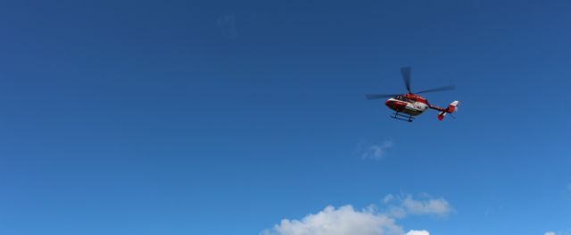 Helicopter flying against bright blue sky with small clouds. Clear daylight. Ideal for themes involving aviation, medical emergency services, transportation, and travel. Can be used in news articles, educational materials, or marketing promotions for aviation or emergency services.