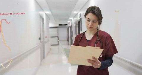Nurse reading patient files in hospital corridor wearing maroon scrubs and stethoscope