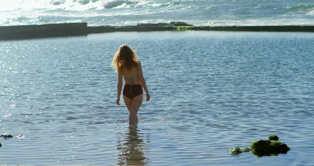 Woman in swimsuit walking into calm ocean water at sunny beach. Ideal for vacation, relaxation, and lifestyle themes. Great for travel brochures, wellness blogs, and summer advertising.