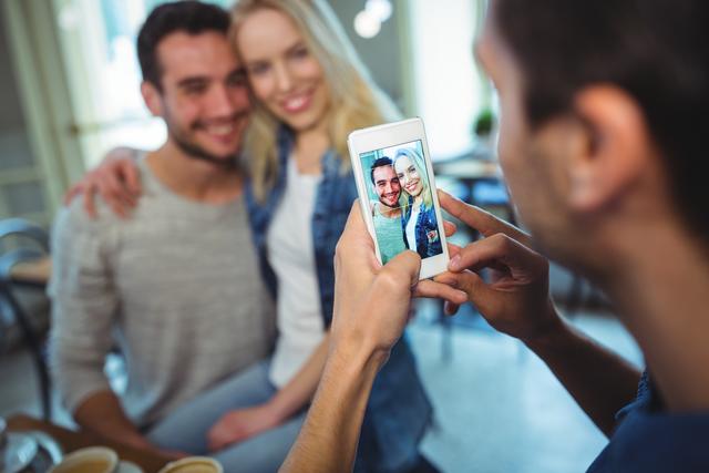 Friend capturing a happy moment of a smiling couple in a café using a mobile phone. Perfect for themes related to friendship, social media, lifestyle, and casual outings. Ideal for use in blogs, social media posts, advertisements, and articles about modern relationships and technology.