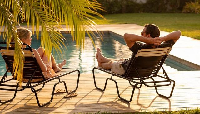 Couple relaxing on lounge chairs by sunlit backyard pool at golden hour
