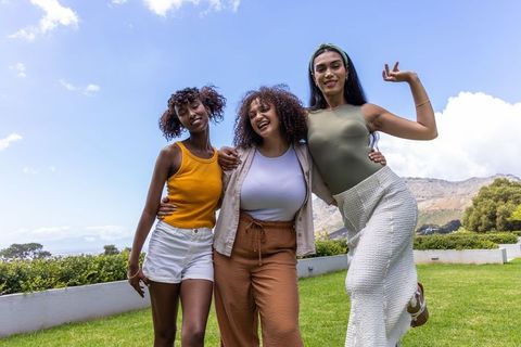 Laughing diverse women linking arms on sunny coastal lawn with hills and blue sky