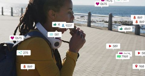Woman eating snack on seaside boardwalk with headphones, backpack and social media alerts