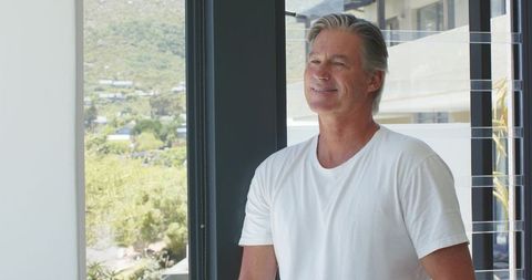 Mature man enjoying scenic hill view from modern glass balcony in bright minimalist home