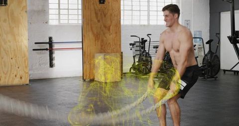 Shirtless man powering battle ropes in industrial gym for high-intensity cardio workout