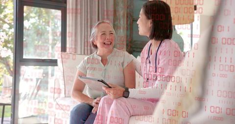 Nurse in pink scrubs showing tablet to laughing senior woman on living room sofa