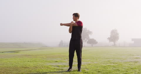 A person is performing stretching exercises on a foggy cricket field, suggesting early morning preparation routine. The mist adds a calm, serene atmosphere to the scene. Suitable for concepts related to sports training, fitness routines, wellness practices, dedication, and outdoor exercises.