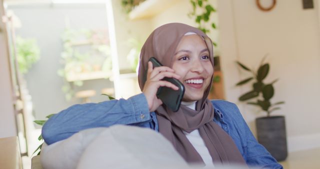 Image of smiling biracial woman in hijab talking on smartphone relaxing in living room at home. Happiness, communication, relaxation, inclusivity and domestic life.