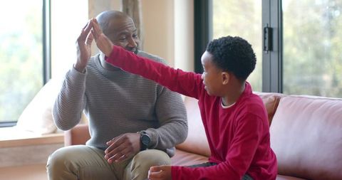 African american older man and young boy giving high-five on cozy living room couch