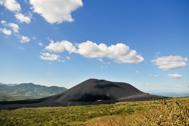 Black volcanic mountain stands prominently under clear blue sky with scattered white clouds. Grassland stretches in foreground providing contrast to the dark volcanic surface. Suitable for use in travel promotions, educational materials, nature documentaries, or outdoor adventure advertisements.
