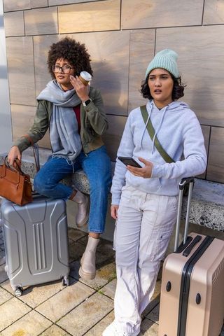 Two women waiting at urban transit bench with luggage, holding coffee and checking phone