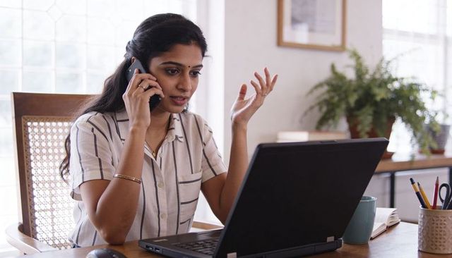 Indian woman working from home on laptop while talking on phone and gesturing