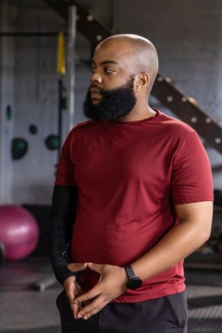 Bearded man standing in gym wearing maroon shirt and smartwatch, focused athlete portrait