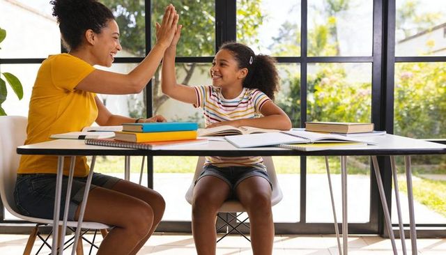 Mother and daughter high-fiving while studying together at home table with books