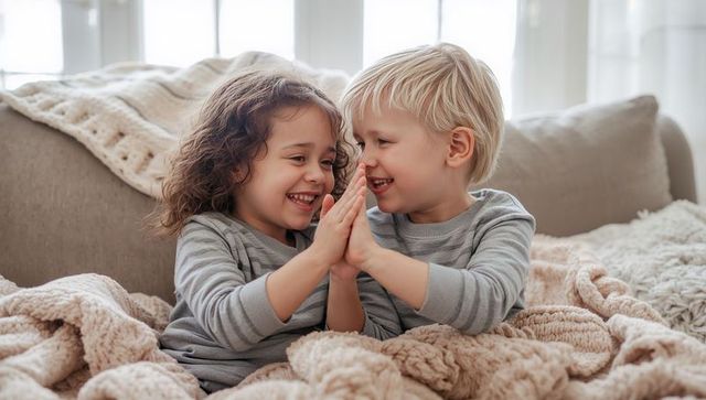 Smiling siblings clapping together on cozy sofa with chunky blankets and cushions, lifestyle