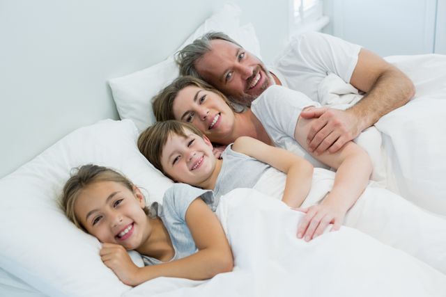This image shows a happy family of four lying together on a bed in a bedroom. The parents and children are smiling and appear to be enjoying a moment of togetherness and bonding. The setting is cozy with white bedding, creating a warm and relaxed atmosphere. This image can be used for promoting family values, home life, and products related to bedding or family activities.