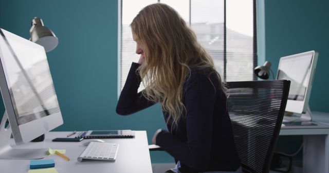 Young woman working at her desk in a modern, blue-colored office. She is focused on a desktop computer, with notes and various stationery surrounding her. Perfect for corporate presentations, articles on women in tech, productivity blogs, modern workplace design inspirations, and stock images for business and technology.