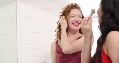Smiling friends applying makeup at bathroom mirror sharing beauty routine