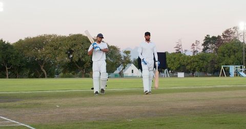 Diverse male cricket batsmen walking on prepared pitch during golden hour at club ground