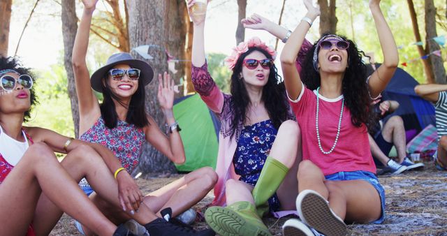 Three young women sitting on the ground in casual festival clothing, raising hands and cheering. They are wearing vibrant outfits, sunglasses, and smiling under the natural canopy of trees. Ideal for themes about friendship, enjoyment, outdoor activities, summer festivals, youthful energy, and celebration.