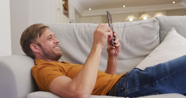 A man is lounging comfortably on a couch while using a digital tablet. He is smiling and appears relaxed in a casual home setting. Perfect for illustrating concepts related to home comfort, leisure time, technology use, or a relaxed lifestyle. Suitable for advertisements, articles, and blogs focusing on home living, digital lifestyles, or relaxation.